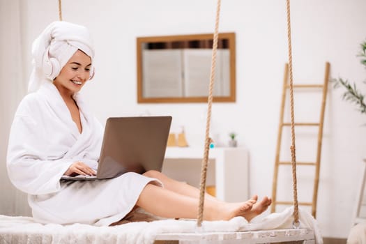 woman in bathrobe and towel after shower uses laptop in bedroom