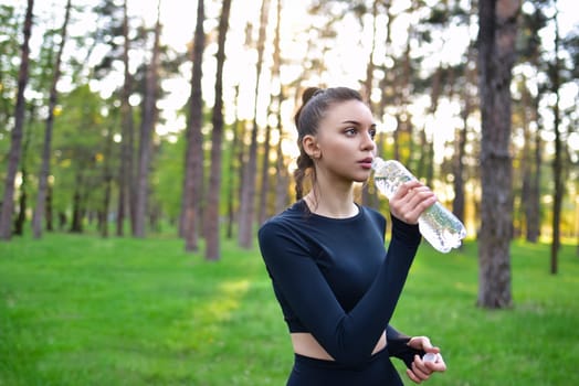 Young woman in sporty black top drinking water in nature from a bottle after jogging in the forest