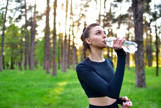 A woman in a sporty black top holds up a water bottle for a drink of water after a sport in the forest