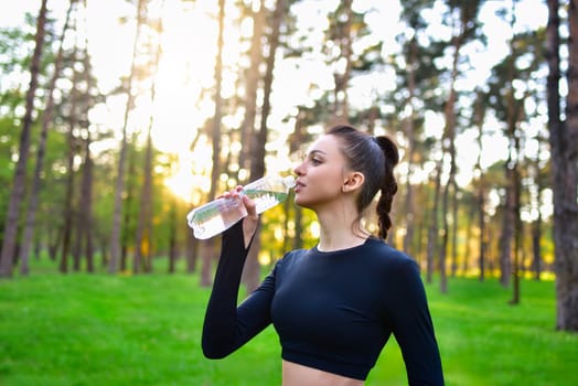 A woman in a sporty black top holds up a water bottle for a drink of water after a sport in the forest
