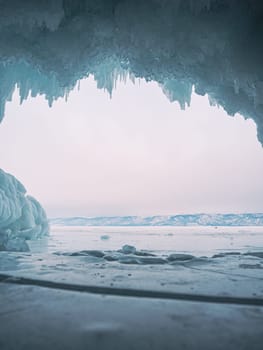 Inside a stunning ice cave on Lake Baikal, large icicles hang from the ceiling, creating a breathtaking winter landscape. Snow-covered mountains can be seen far in the distance.