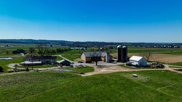 Overhead shot of a busy farm complex, featuring multiple barns and silos surrounded by verdant pastures, equipment in use, and a group of people gathered, under a clear sky.