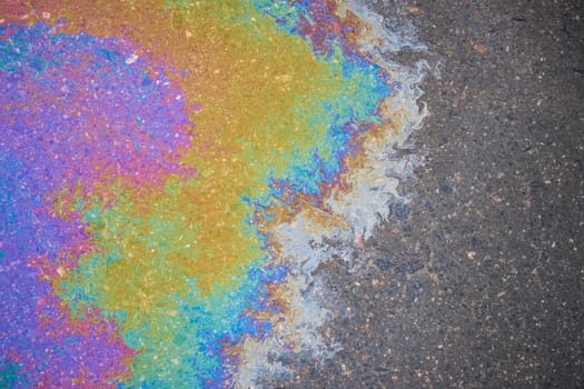 Residues of oil on asphalt left by rain form trails that scatter sunlight, creating a rainbow effect