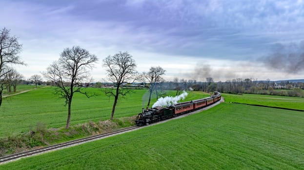 Dramatic aerial capture of a vintage steam train chugging along a curvy track through lush green fields, with a backdrop of dramatic cloudy skies and a distant village.