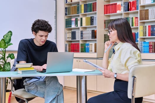 Young male college student talking studying with female teacher. Mentor and guy together in educational library office room. Education, training, mentoring, teaching, youth concept