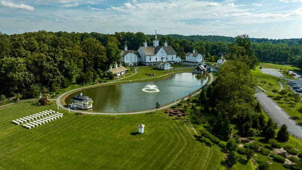 Aerial View Showcasing A Cluster Of Traditional White Orthodox Churches With Cross-Topped Domes, Arranged Around A Curved Pond With A Fountain, Amidst Green Trees And Neatly Arranged White Benches