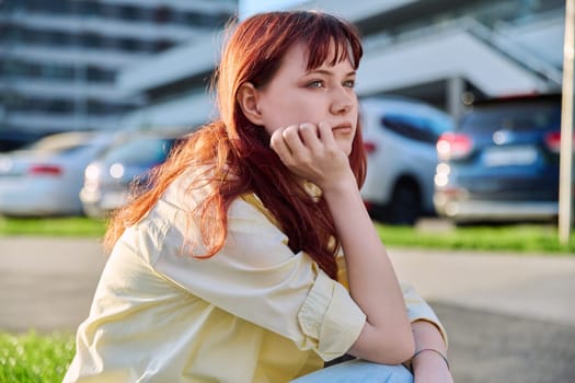 Upset sad unhappy young female sitting on steps. Pensive serious beautiful red-haired girl student 19-20 years old sitting outdoor. Problems, difficulties, depression, mental health of young people