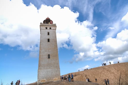 Tourists walk on the sand near the Rubjerg Knude Lighthouse on the coast of the North Sea in the Jutland in northern Denmark. Natural landscape with sand dunes. Rubjerg, Denmark - August 24, 2023.