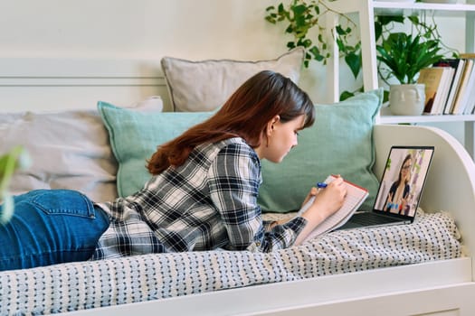 Female student lying on couch at home, looking at laptop computer screen, having remote video lesson, consultation, listening to online course, writing in notebook. Technology education study youth