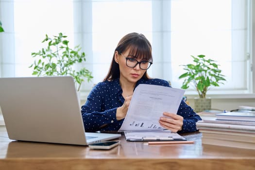 Middle-aged serious woman working with laptop computer with business papers, sitting at desk in home office interior. Work, remote business, freelance, technology, 40s people concept