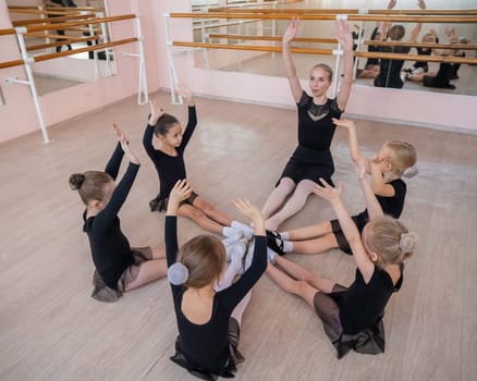 Caucasian woman and five little girls sit in a circle and do stretching at a ballet school