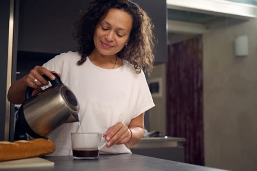 Multi ethnic smiling woman in white t-shirt making coffee at home, pouring boiled water from a teapot into a glass cup, standing at kitchen table with a loaf of wholesome bread on the cutting board