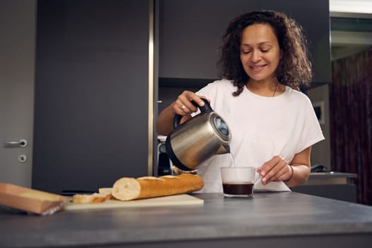 Smiling Latin American woman at home, making coffee. Pretty brunette n white pajamas, standing at kitchen table, preparing breakfast, poring water from a teapot into a glass and making sandwiches