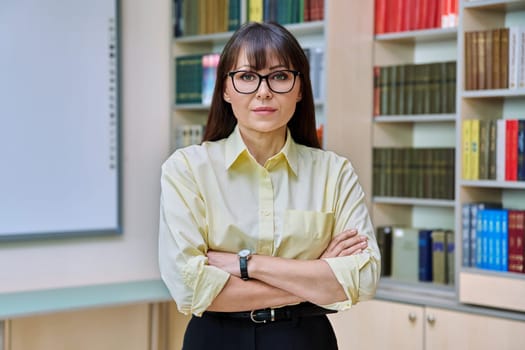 Portrait of confident mature business woman in library. Middle-aged elegant female with crossed arms, manager teacher mentor psychologist counselor advisor librarian background of shelves with books