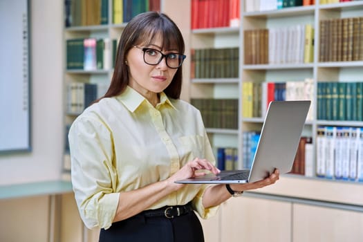 Middle aged elegant woman using laptop computer inside library. Confident serious elegant business female looking at camera teacher mentor professor leader manager psychologist counselor social worker