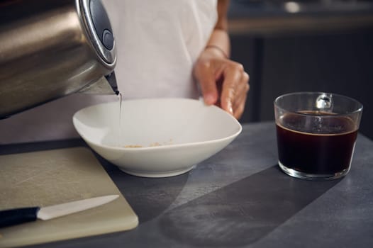 Close-up woman pouring boiled water from a stainless steel teapot into a white bowl with oat flakes, preparing dry breakfast, standing t kitchen counter at home. Food and drink concept. Healthy eating