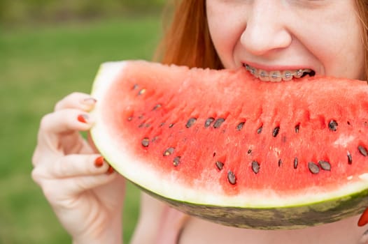 Close-up portrait of red-haired young woman with braces eating watermelon outdoors.