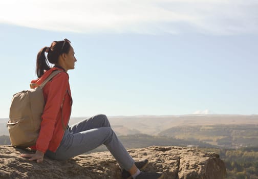 A girl tourist with a backpack sits on the top of a mountain and enjoys the beautiful views. A walking woman in an orange jacket rests on a rock, looking at the beautiful sunny landscape of the Caucasus Mountains