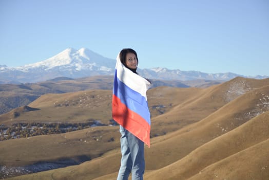 A young brunette woman stands against the backdrop of the snow-capped Mount Elbrus, looking at the camera, a Russian flag covers her shoulders. Tricolor against the backdrop of snow-capped Mount Elbrus.