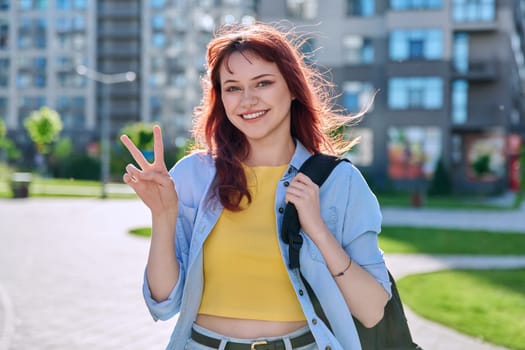 Young smiling happy beautiful red-haired hipster female looking at camera showing victory peace gesture with her hand, outdoor in sunlight. Beauty, fashion, style, lifestyle youth concept