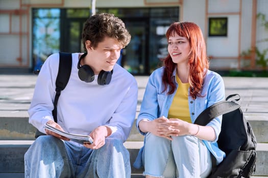 Teenage college students guy and girl talking, sitting outdoor near educational building. Youth 19-20 years old, education, lifestyle, friendship concept