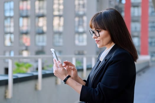 Mature business confident female using smartphone, modern city background. Middle aged successful woman texting using mobile applications apps for business work. Technology internet job, urban style