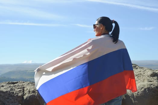 A girl tourist with a Russian flag on her shoulders stands on the top of a mountain and enjoys the beautiful views of the Caucasus Mountains