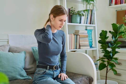 Young teenage female experiencing neck pain sitting on couch at home. Symptoms of the musculoskeletal system, neurological pathologies, acute infectious diseases