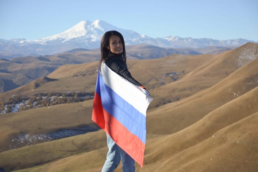 A young brunette woman stands against the backdrop of the snow-capped Mount Elbrus, looking at the camera, a Russian flag covers her shoulders. Tricolor against the backdrop of snow-capped Mount Elbrus.