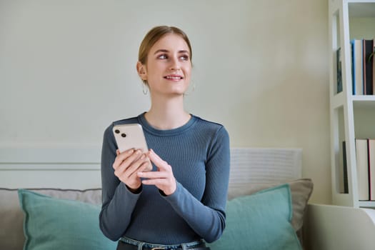 Happy smiling female teenager using smartphone, sitting on couch at home, girl 16,17, 18 years old texting reading messages. Modern digital technologies for communication, leisure, learning, shopping