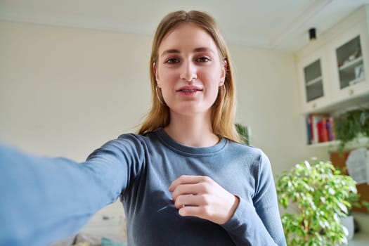 Close-up selfie portrait of teenage cheerful grimacing young female looking at web camera. Happy teenage girl 16, 17, 18 years old with blond hair, in home interior. Beauty, lifestyle, youth concept