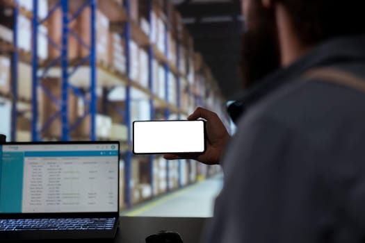 Storehouse employee looking at phone with blank display template, verifying industrial stock labels and order receipts. Supervisor providing cargo merchandise for supply chain contracts.
