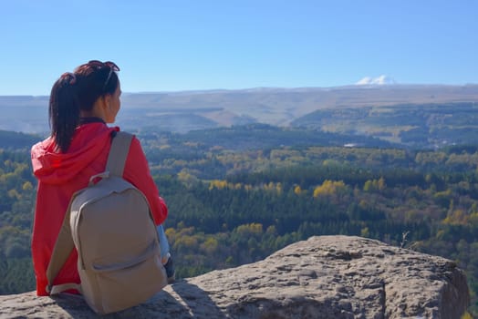 A girl tourist with a backpack sits on the top of a mountain and enjoys the beautiful views. A walking woman in an orange jacket rests on a rock, looking at the beautiful sunny landscape of the Caucasus Mountains