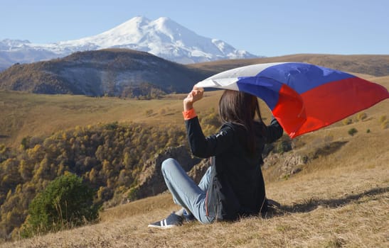 A young brunette woman sits on a stone and looks at the Caucasus Mountains around, a Russian flag flutters in her hands. Tricolor against the backdrop of snow-capped Mount Elbrus.