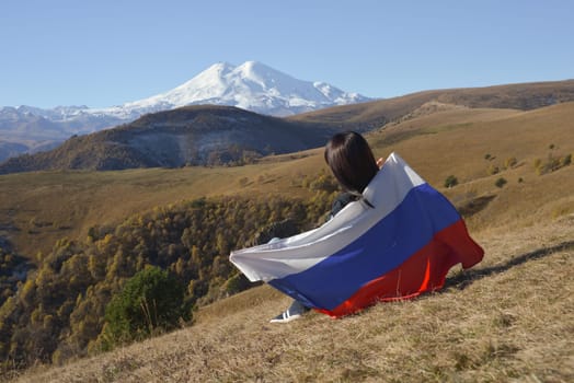 A young brunette woman sits on a stone and looks at the mountains around, her shoulders are covered with a Russian flag. Against the backdrop of snow-capped Mount Elbrus.