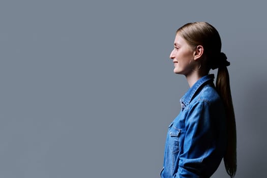 Profile view, young smiling female looking forward on gray studio background, copy space for advertising text image. Beautiful teenage blonde with long hair in denim shirt, fashion style beauty youth