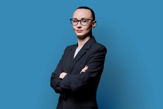 Portrait of business woman in 30s with crossed arms, on blue studio background. Confident female in glasses, suit looking at camera. Business, work, job, career, people concept