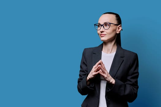 Portrait of thinking serious 30s business woman on blue studio background. Confident female in glasses suit looking to side at space for image text copy space. Business work teaching job career people