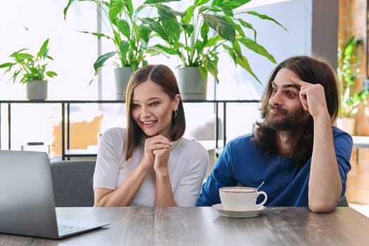 Happy smiling surprised young couple looking at laptop together while sitting in cafe, cafeteria. Leisure time for two, lifestyle, togetherness, relationship, communication, work study remotely