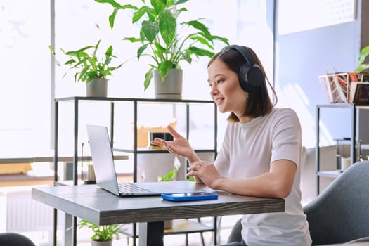 Young woman in headphones having video chat conference call using laptop computer sitting in workshop cafe. Female college student studying online listening webinar preparing exam, working remotely