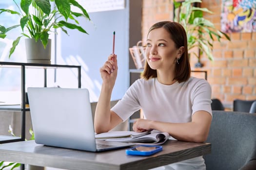 Young female college student studying using laptop, writing in notebook while sitting in coworking cafe. E-learning, education, lifestyle, youth concept