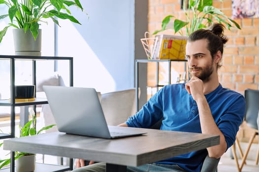 Young serious man working and studying using laptop sitting in coworking cafe. 30s handsome bearded guy looking at computer screen. Technology for leisure work business education communication people