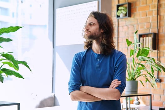 Portrait of young serious confident handsome man with long hair and beard, with crossed arms, profile view, copy space