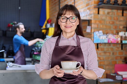 Woman in apron, food service coffee shop worker, small business owner with cup of fresh coffee, looking at camera near bar counter with male barista. Staff, occupation, entrepreneur, work concept