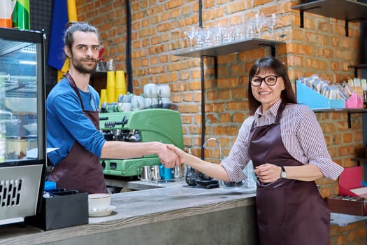 Young man in apron shaking hands with woman owner of coffee shop cafeteria restaurant, colleagues partners working together. Cooperation, small business, partnership, teamwork team, staff, success