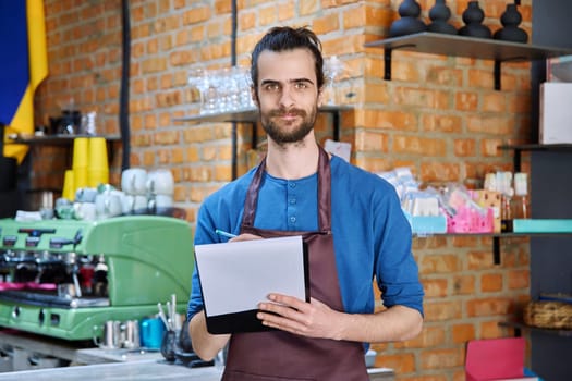 Young man in apron, food service worker, small business owner entrepreneur with work papers looking at camera near counter of coffee shop cafe cafeteria. Staff, occupation, successful business, work