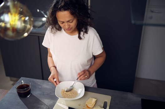 Top view curly haired multi ethnic middle aged woman preparing dry breakfast at home, holding a spoon and mixing oat flakes with milk, standing at kitchen counter in minimalist home kitchen interior