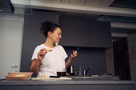 Authentic young adult woman 40s, eating sandwich with cheese and holding her smartphone in hands, smiling looking aside, standing at kitchen counter in minimalist home kitchen interior.