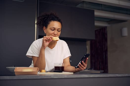 Beautiful multi ethnic woman 40s, eating sandwich with cheese and checking social media content on his smartphone, standing at kitchen counter in minimalist home kitchen interior.