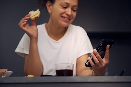 Details on a modern mobile smartphone in hands of a smiling woman, checking social media content, browsing web, scrolling newsfeed, standing at kitchen counter with a sandwich in hand at home